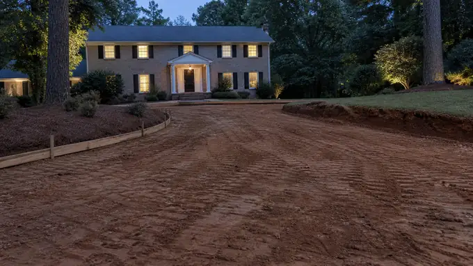 A plate compactor tamps down soil in a small excavated area. at a tan-brick colonial in Alpharetta, GA, at blue hour.